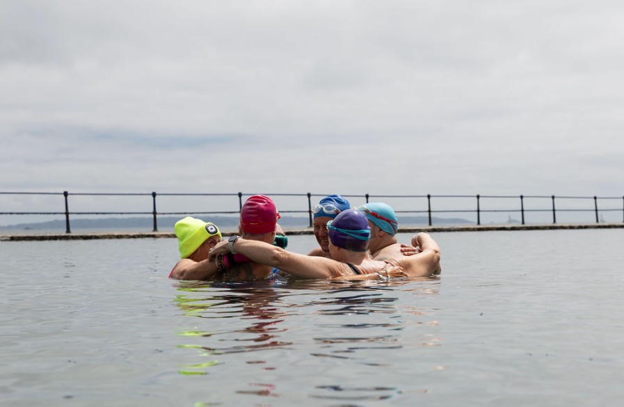 A group of sea swimmers stand together in the water in an outdoor tidal pool, with their arms around each other.