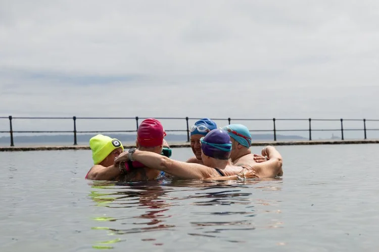 A group of sea swimmers stand together in the water in an outdoor tidal pool, with their arms around each other.