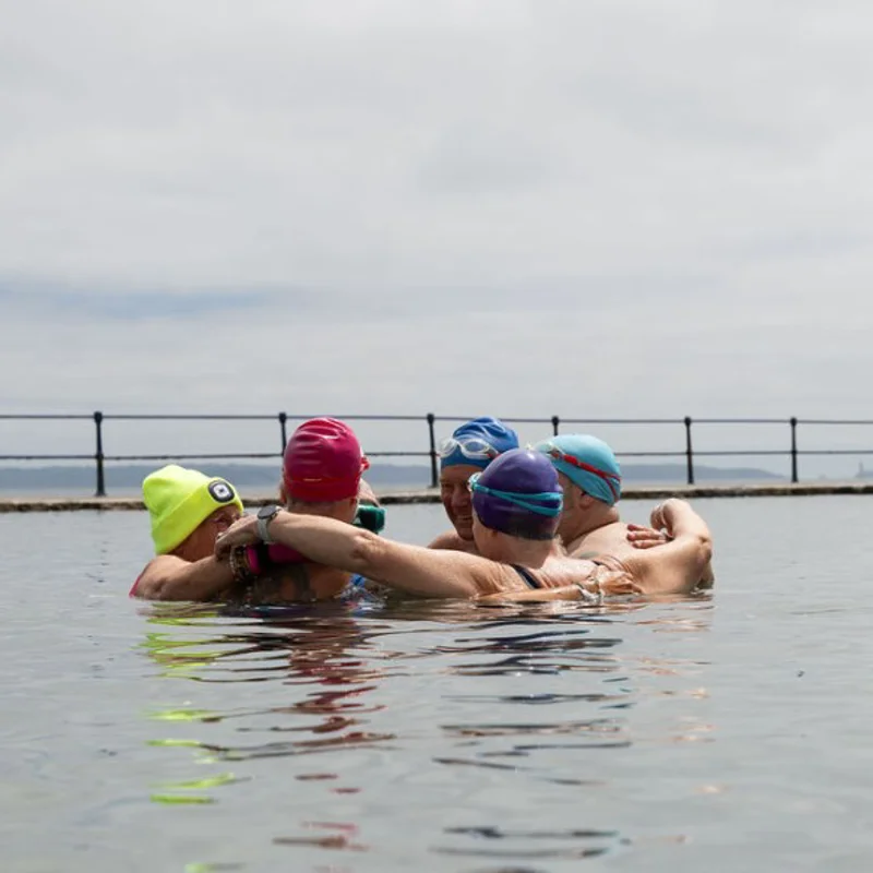 A group of sea swimmers stand together in the water in an outdoor tidal pool, with their arms around each other.