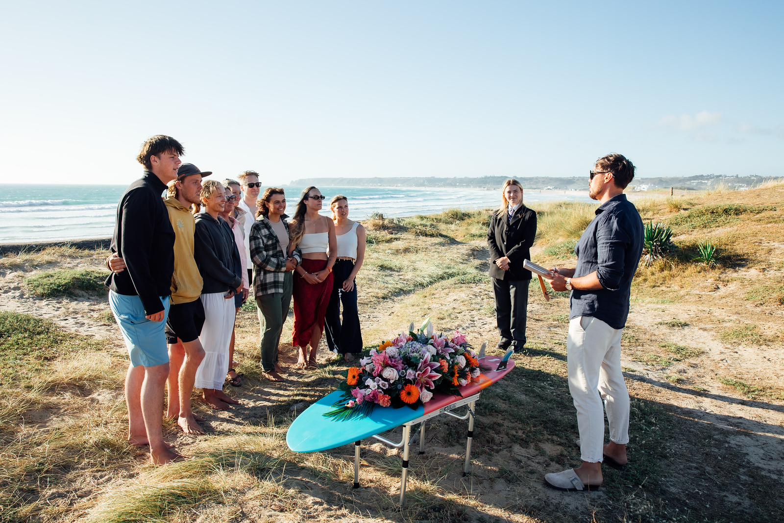 Family and friends gather on the beach for a memorial ceremony led by a celebrant, with flowers arranged on a surfboard.