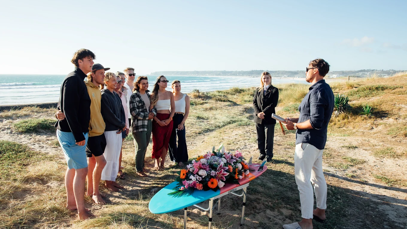 Family and friends gather on the beach for a memorial ceremony led by a celebrant, with flowers arranged on a surfboard.