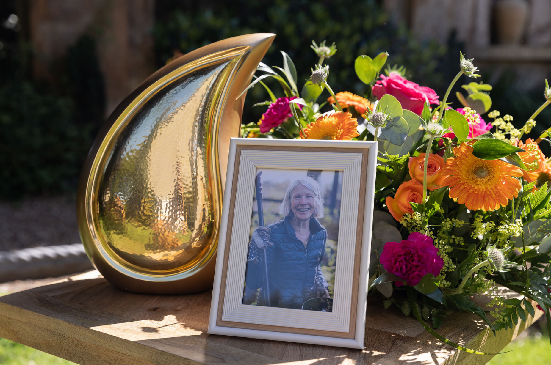 Memorial display featuring a decorative urn, framed photograph and colourful floral arrangement on a wooden table.