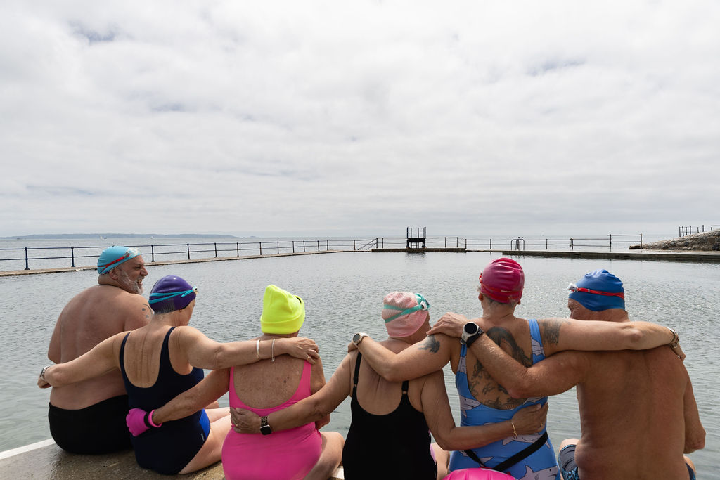 A group of sea swimmers sit together by an outdoor tidal pool, with their arms around each other.
