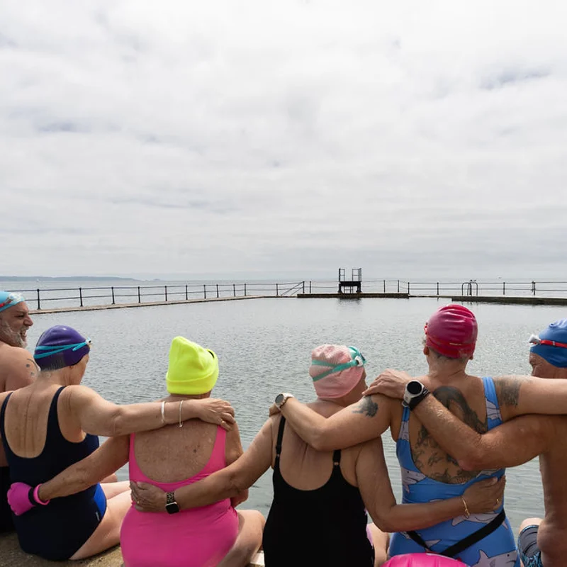 A group of sea swimmers sit together by an outdoor tidal pool, with their arms around each other.