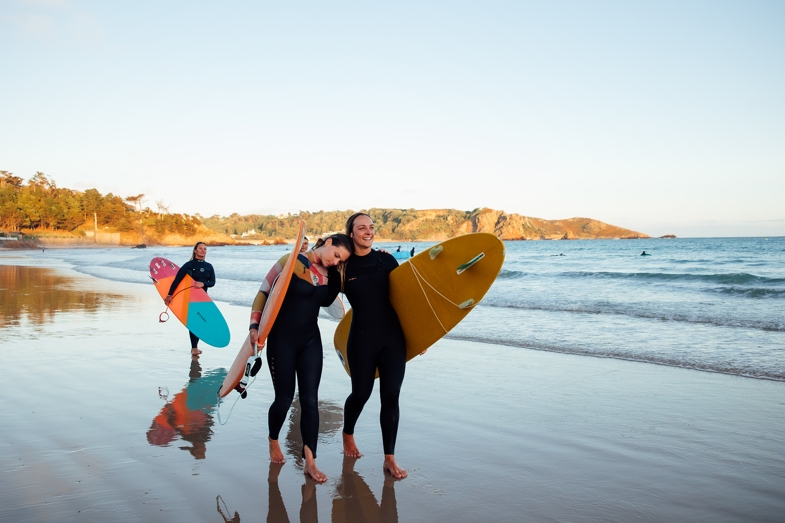 Two surfers walk along the beach carrying surfboards, with another surfer behind them.