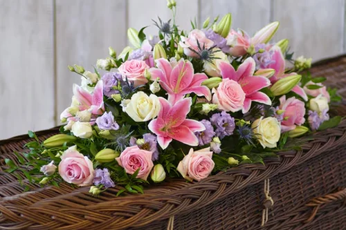 Funeral flower arrangement with pink lilies, roses and complementary blooms displayed on a wicker coffin.