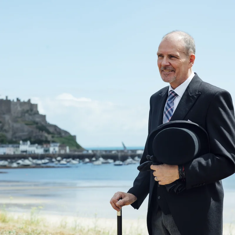 Funeral director from Coop Funeral Care by De Gruchy in traditional funeral morning dress standing in front of Gorey Castle overlooking Grouville Bay.