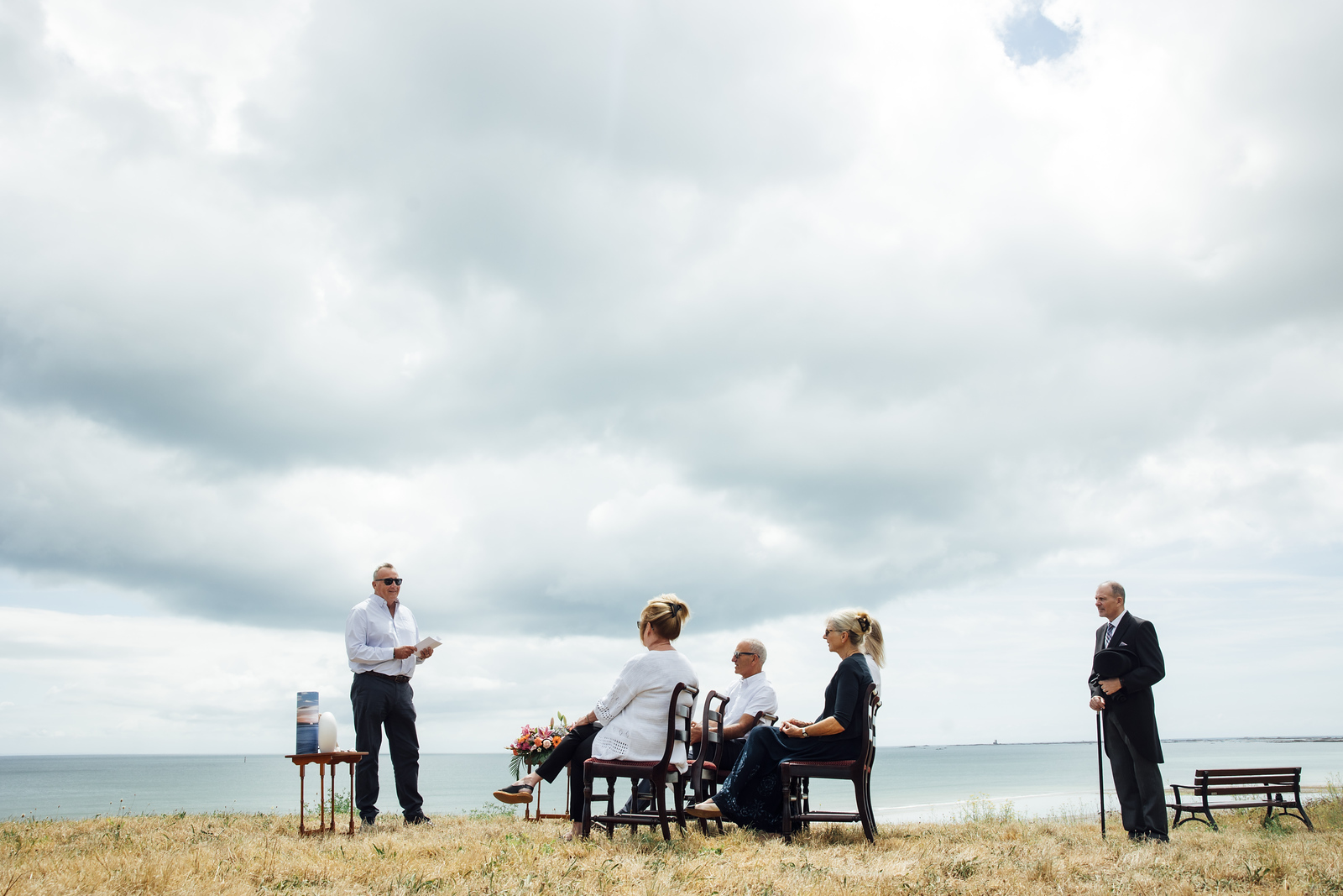 Family members sit by the sea during a memorial service led by a celebrant, with a funeral director standing nearby.